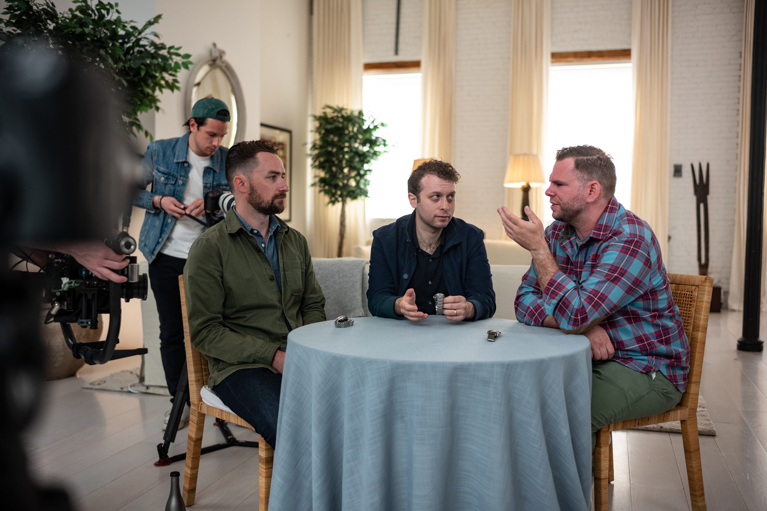三个男人围坐在一张桌子旁，与背景中的摄影师交谈